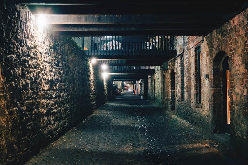 Dark and eerie lower-level alley at night surrounded by brick walls on two sides and old iron crosswalks above