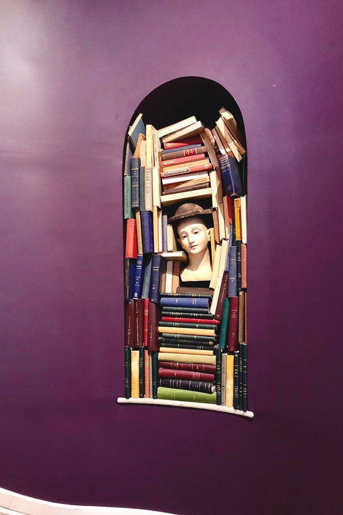 An oval wall niche stuffed with books and a mannequin head, inset against a deep purple wall