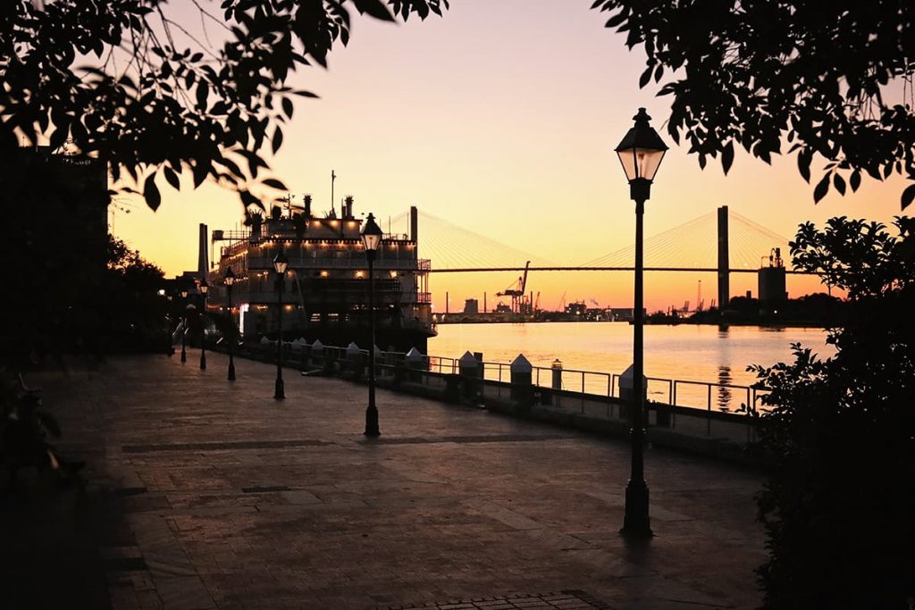 Boat docked by river with yellow and purple sunset in background and dark tree leaves framing the foreground