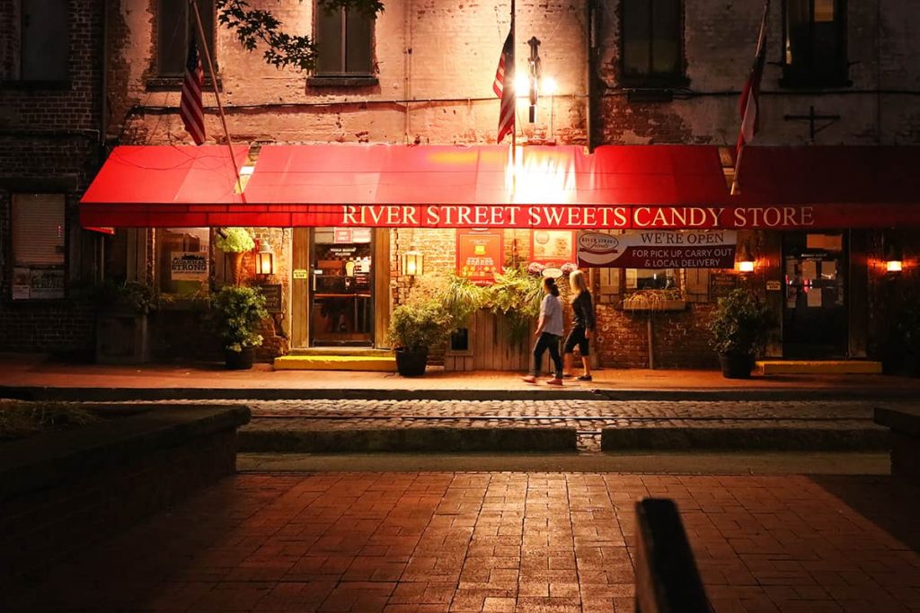 Candy shop with a red awning dimly lit at night with two women passing by