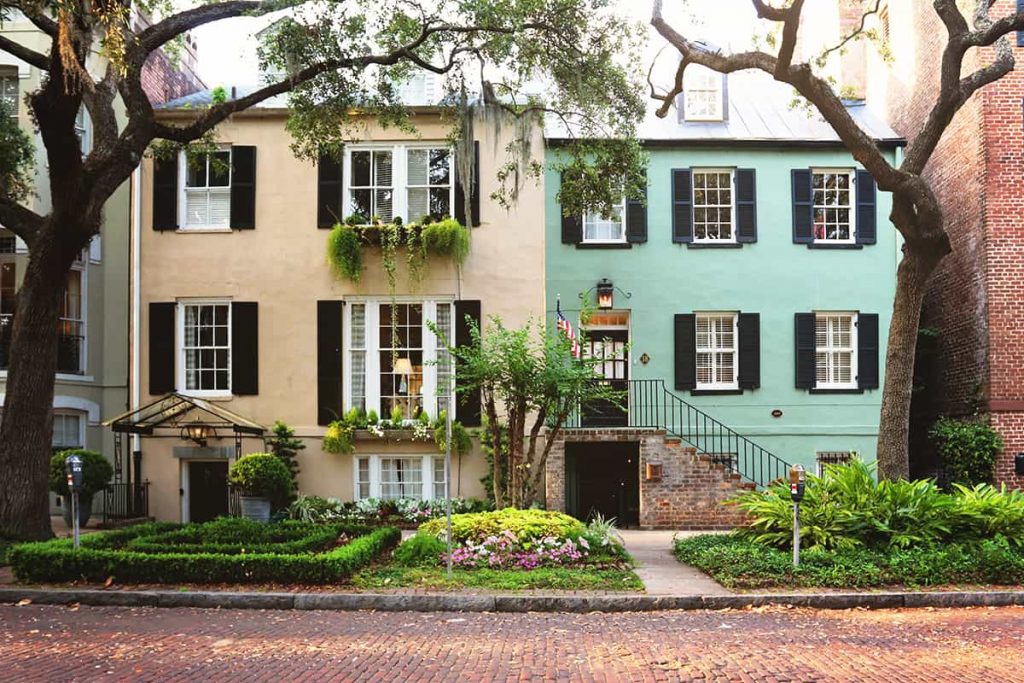 Two attached homes on a brick-paved street with oak trees in the front yard. Tan home to the left and turquoise home on the right