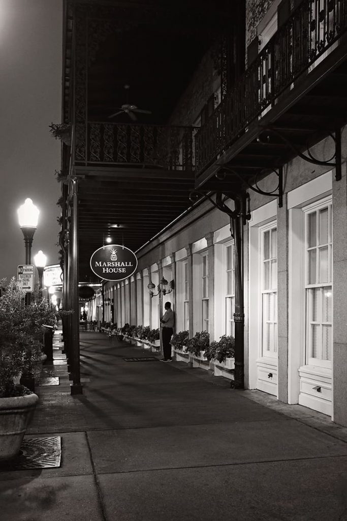 Nighttime view of the front facade of The Marshall House Savannah with an empty streetlit sidewalk and one lone bellhop by the door