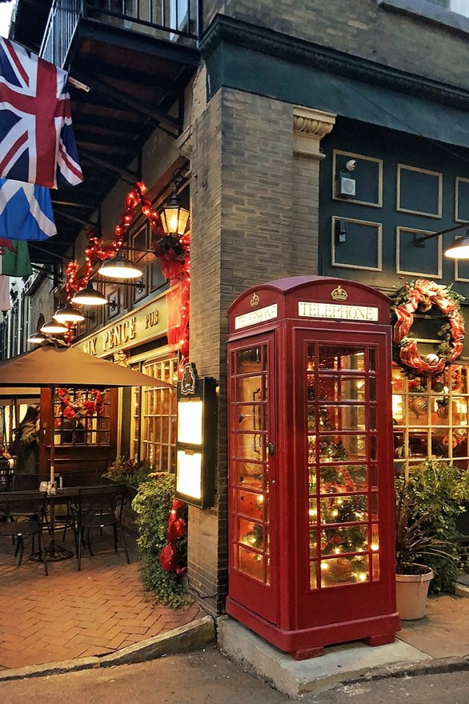 Six Pence Pub with a lit Christmas tree inside a red telephone booth, a wreath over a window, and red ribbons and colorful flags hanging over the front entrance