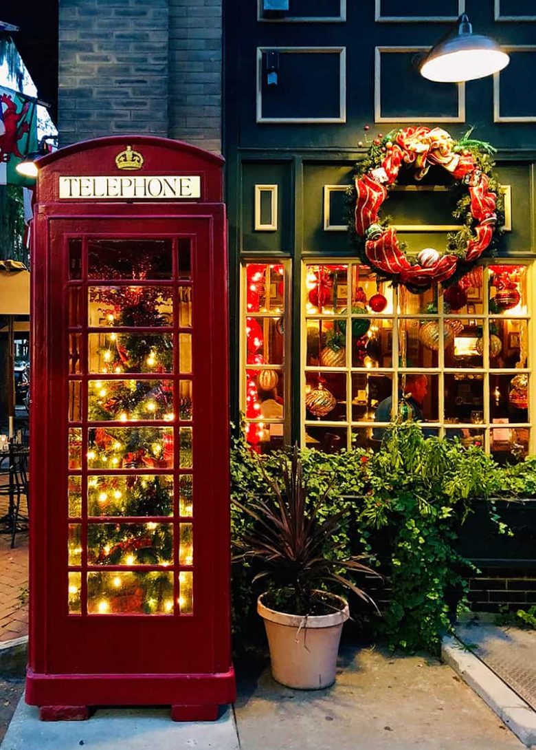 Six Pence Pub with a lit Christmas tree inside a read telephone booth, a wreath hung over a window, and ornaments hanging fro, the interior of the windows