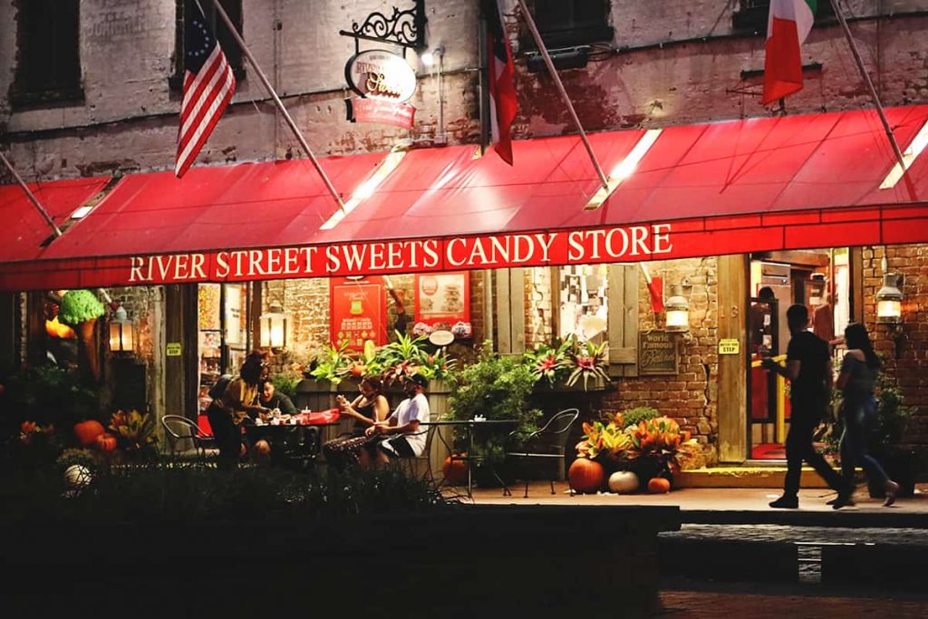 Exterior of River Street Sweets with the red awning lit up at night and pumpkins and fall decor by the door