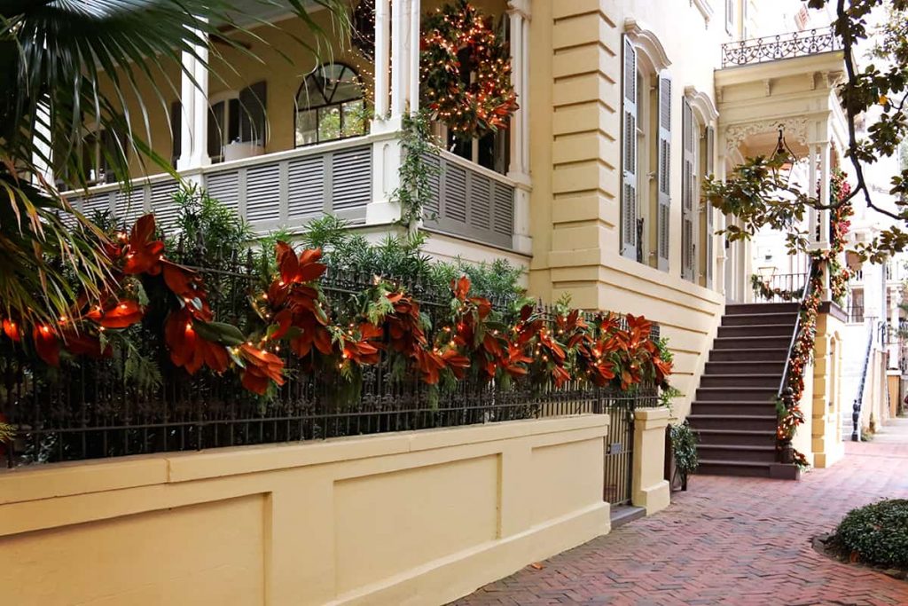 Yellow historic home with a large side porch and magnolia leaf Christmas decorations on the wrought iron fence