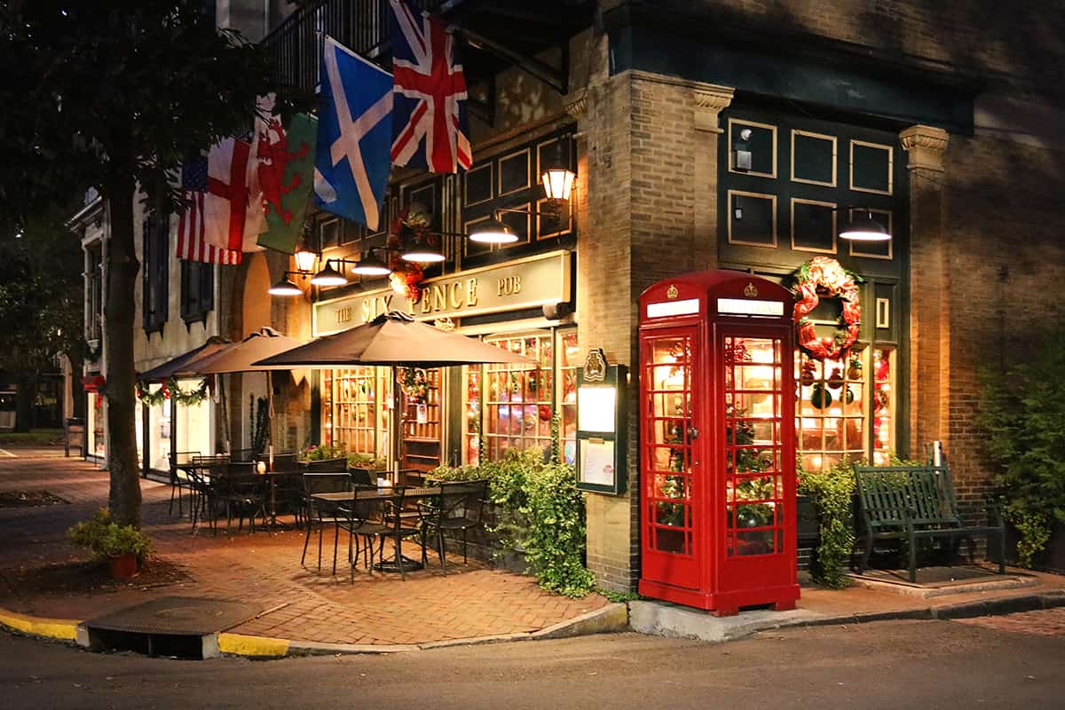 Six Pence Pub storefront in Savannah Georgia with a magnolia tree growing out of the historic brick sidewalk and a bright red phone booth to the right of the door