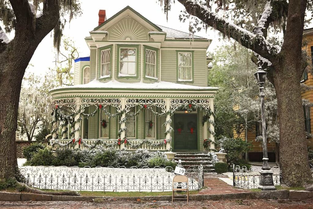 Street view of a well-maintained Victorian-style home painted sage green with dark green trim and a dark green door. The home is decorated for the holidays with greenery and red ribbon and two wreaths on the front doors. There is snow on the roof, ground, and in the large oak trees towering over the home