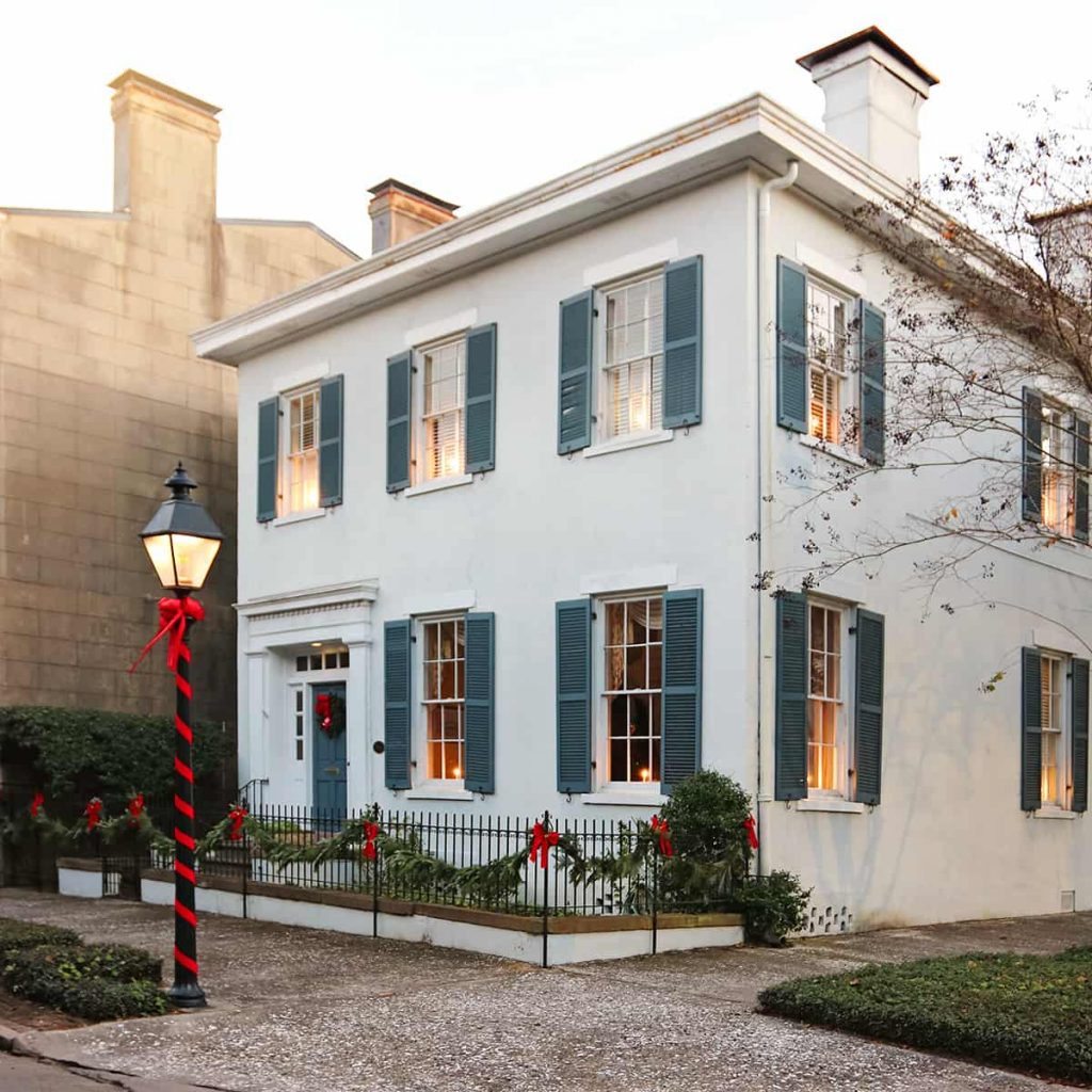 Pale blue historic home with dark blue shutters, candles illuminating the windows, and Christmas greenery on its wrought-iron fence