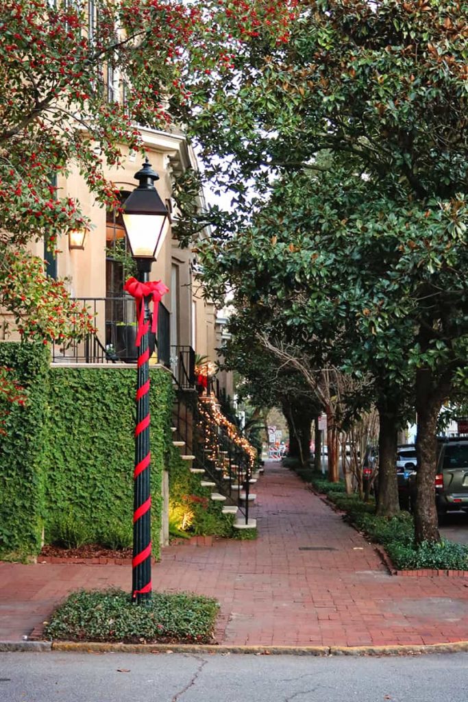 Black streetlamp wrapped in red ribbon with a red bow at the top and a view down a sidewalk lined with historic homes in Savannah