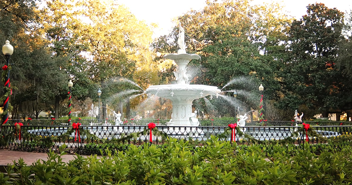 The Forsyth Park fountain illuminated by sunlight with greenery and red Christmas bows decorating the lamp posts