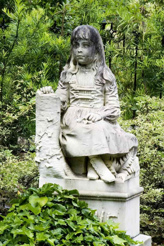 Statue of a little girl with long hair wearing a dress and sitting properly with her ankles crossed