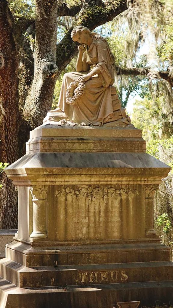 An elaborate funeral marker with Theus carved at the bottom and a woman sitting at the top with a forlorn look on her face
