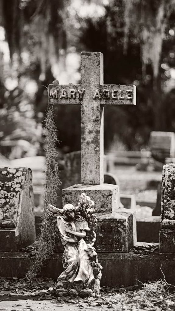 An old weathered stone cross with lichens and Spanish moss growing on it and an angel statue in front of it