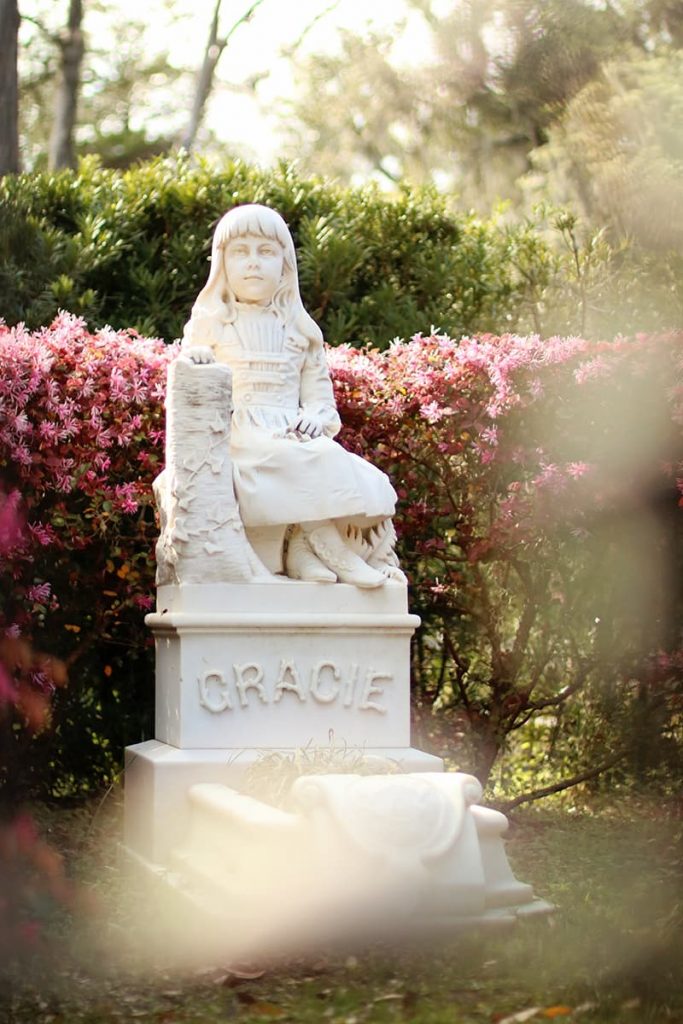 Statue of Little Gracie Watson in Bonaventure Cemetery with pink blooming flowers and greenery behind her