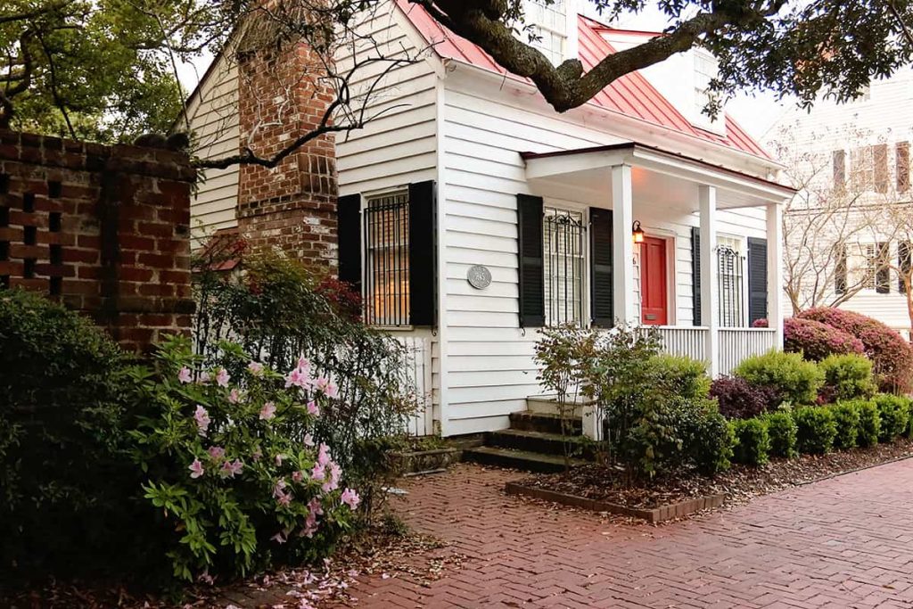 The Henry Willink Cottage, a charming tiny house with white boards, black shutters, and a reddish metal roof. The home is surrounded by beautiful landscaping and brick sidewalks
