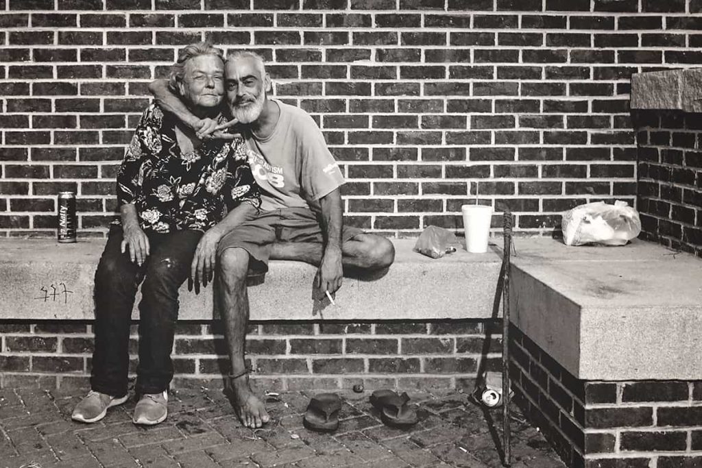 B&W photo of a thin, older man homeless man in Savannah with a gray beard and bare feet. His arm is wrapped protectively around the neck of an older female, and he's throwing a peace sign.