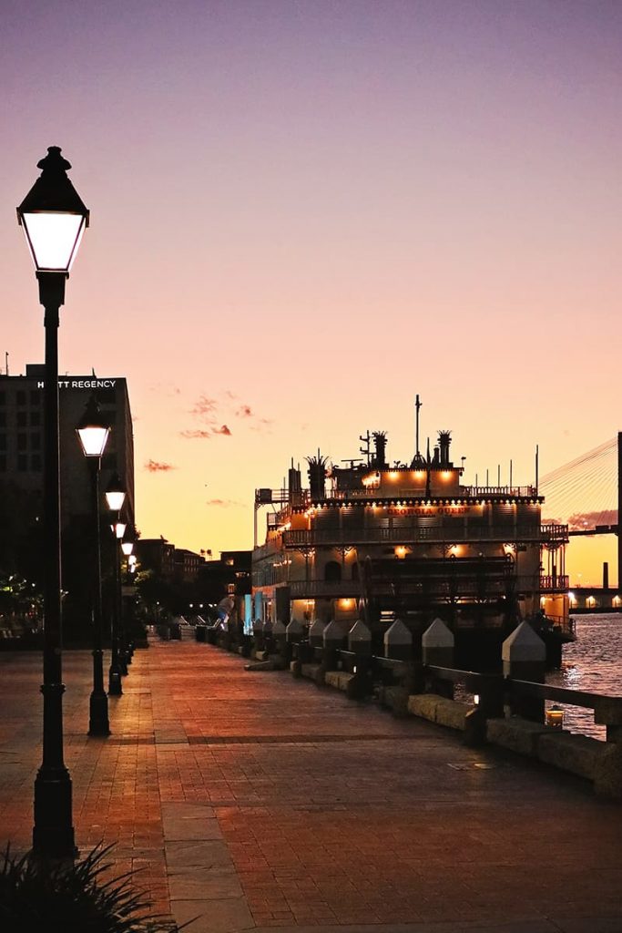 A line of gas lanterns leads the eye down Savannah's River Street towards the Georgia Queen steamboat and a purple, pink, and yellow sunset in the distance