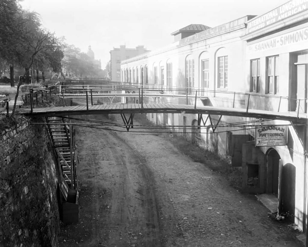 Historic B&W photo of Factors Walk with dirt roads under pedestrian foot bridges along a row of warehouses