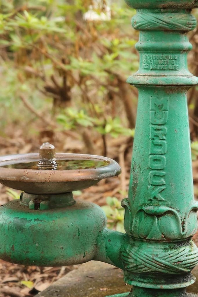 An old and rusted green water fountain with leaf detailing and a maker label that reads Murdock