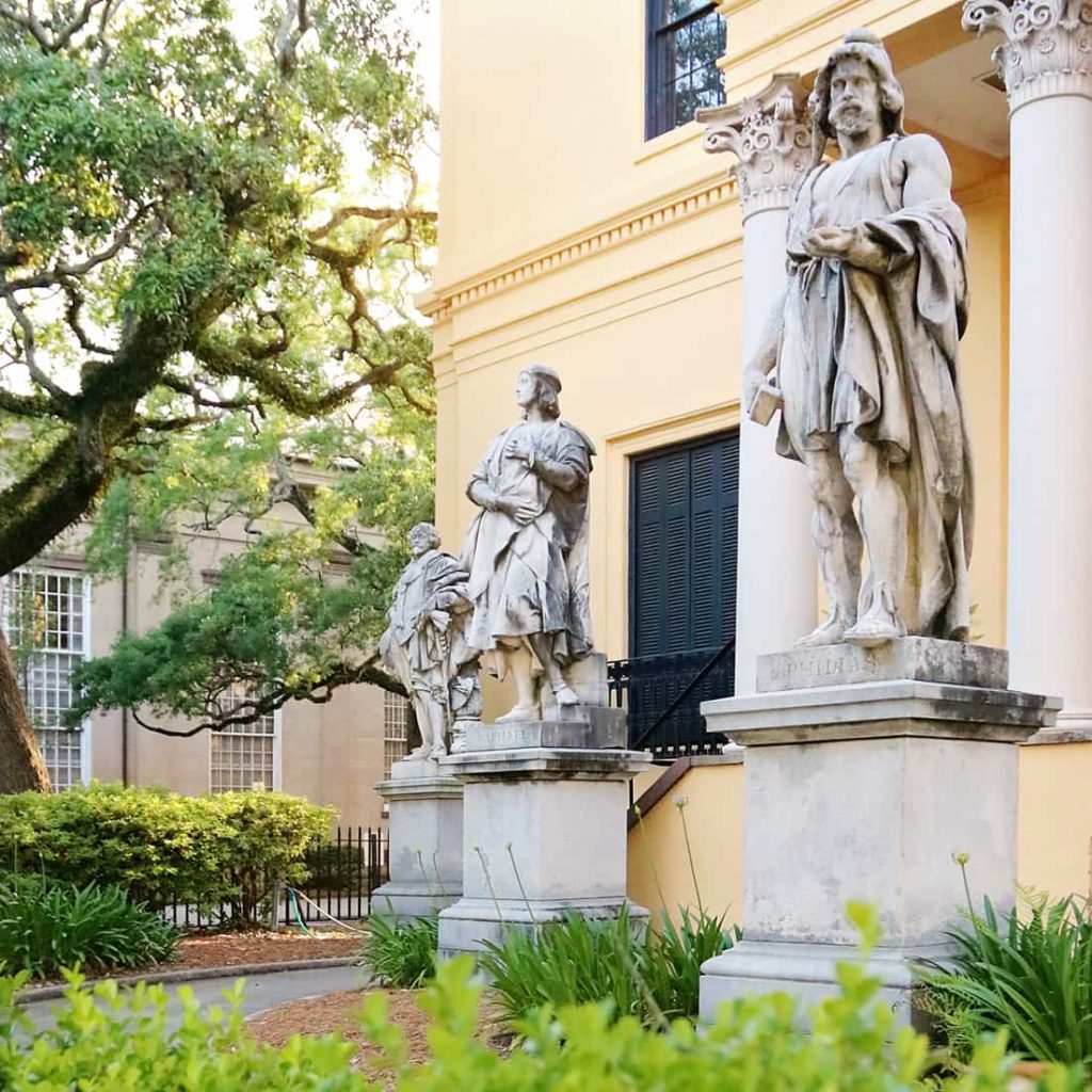 Three statues of men in robed attire stand guard in front of the yellow Telfair Academy building