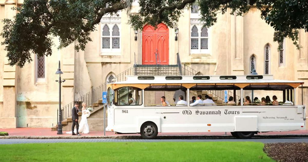 A white trolley with Old Savannah Tours written on its side is parked in front of an old church with red doors. A newly wedded bride and groom walk towards the trolley