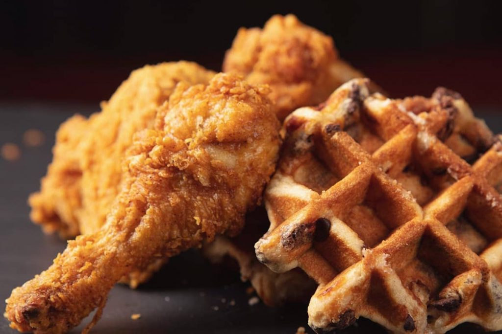 Perfectly crisp, lightly fried chicken legs next to a blueberry waffle on a dark plate background