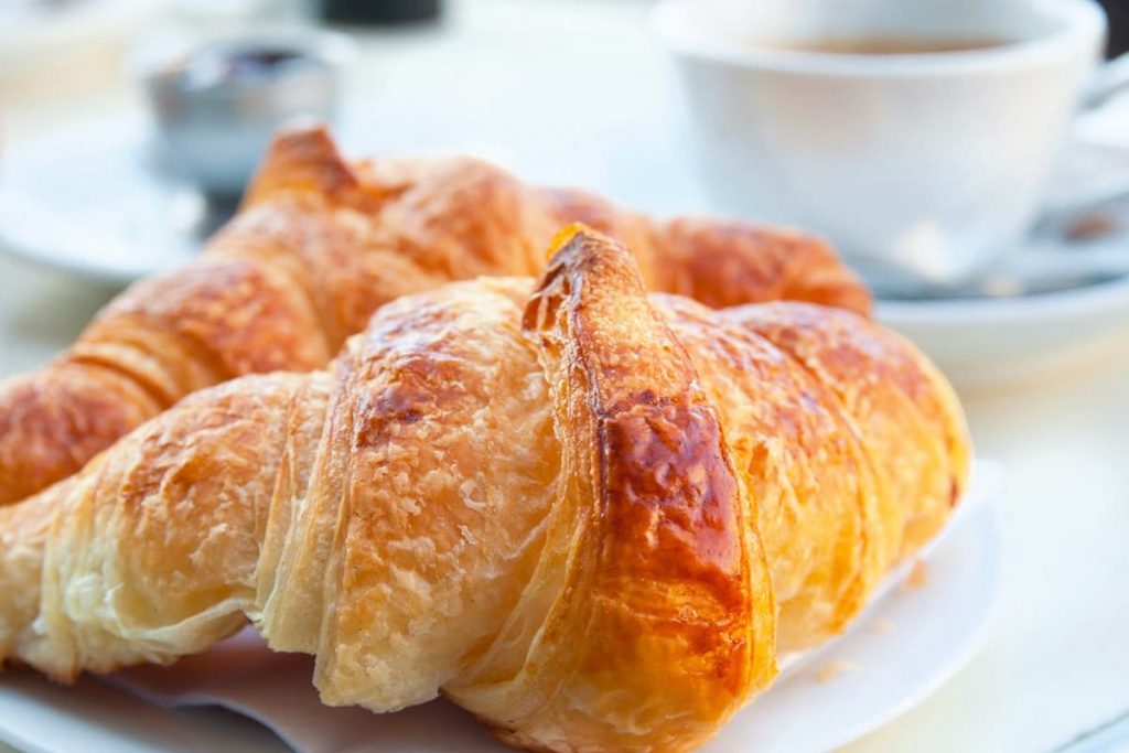 White plate with two flaky, buttery light brown croissants and a white coffee cup in the background