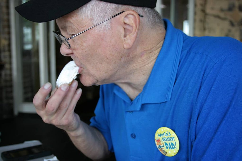 An older gentleman in a blue shirt and black ballcap with a World's Greatest Dad pin on his shirt tastes a beignet at Huey's in Savannah