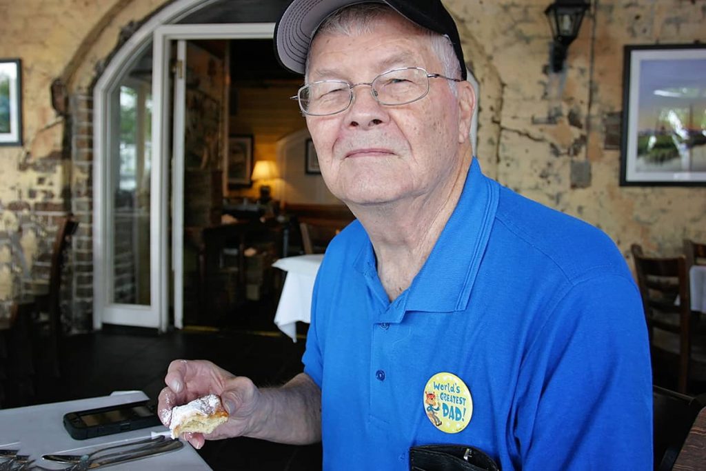 A friendly gentleman in a blue shirt and ball cap smiles as he holds a half-eaten beignet in his hand at Huey's in Savannah, GA