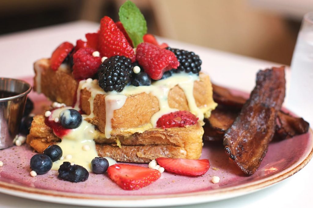 Close up of a pink plate with two pieces of french toast covered in various colorful berries and sweet cream, with a side of bacon at The Collins Quarter in Savannah