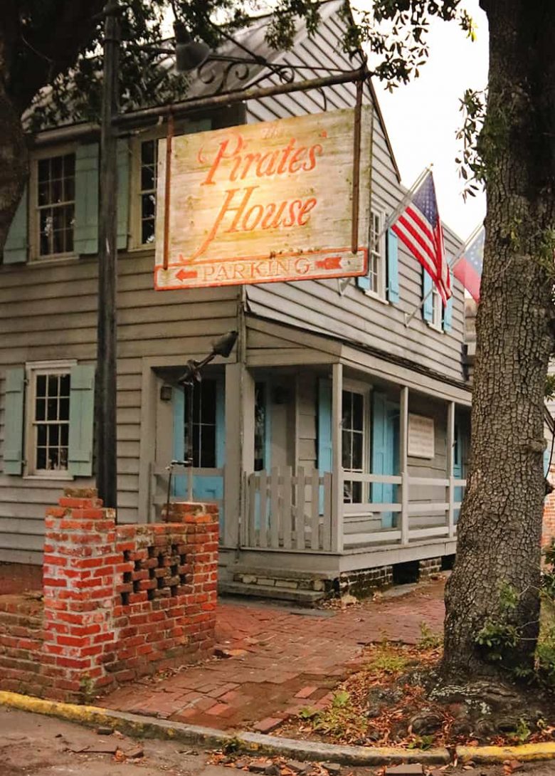 Signage that reads Pirates' House Parking and an old grey wooden inn with haint blue shutters and American flags