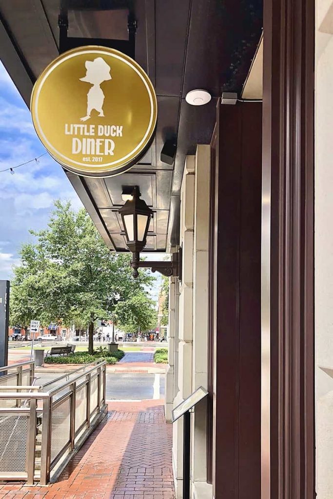 Gold circular sign with a white duck wearing a dapper hat and the words Little Duck Diner written in white