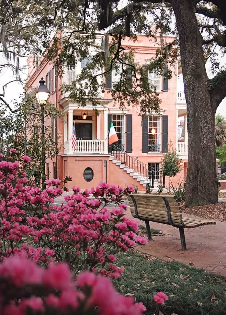 View of 432 Abercorn Street through hot pin azalea bushes in Calhoun Square. The home is a 3-story salmon-colored plaster structure with black shutters and white trim