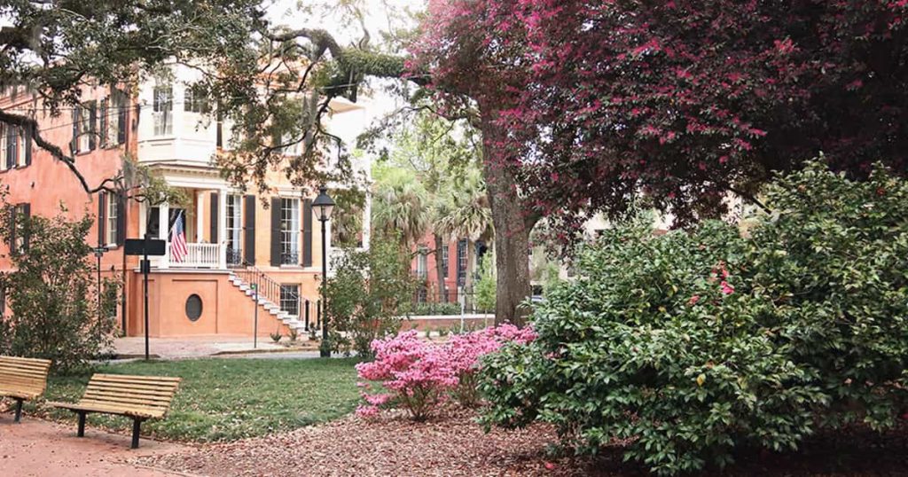 A salmon colored 3-story home in the distance with beautiful landscaping of pink azaleas and large oaks in the foreground