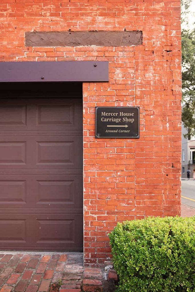 Red brick carriage house with a brown door and sign indicating the entrance to the carriage house at Mercer Williams House