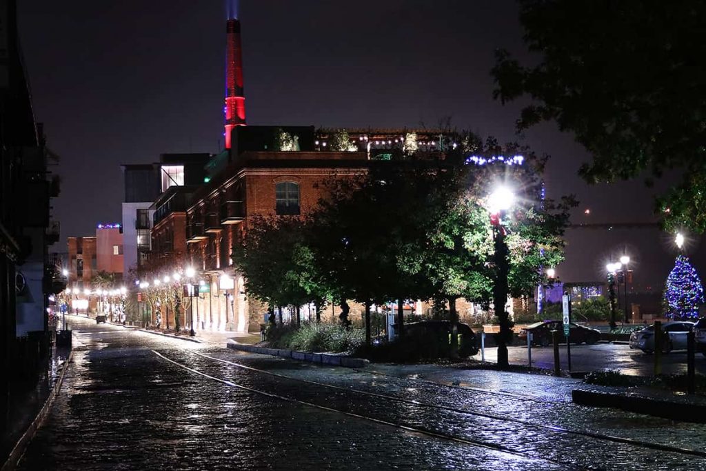 Late-night scene of a the JW Marriott Plant Riverside lit with red and purple lighting which is reflected on wet cobblestone streets in the foreground
