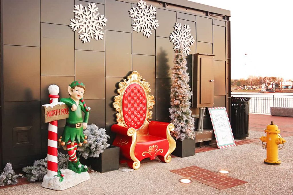 A photo station set up for photos with Santa includes a fancy red and gold chair, a tall skinny Christmas tree, and an elf wearing green and leaning against a sign for the North Pole