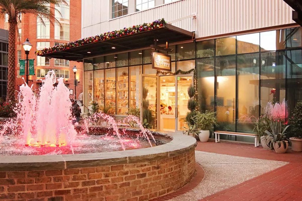 A circular water fountain spouting sprays of water lit with soft pink lighting. A shop in the background is decorated with Christmas ornaments and greenery
