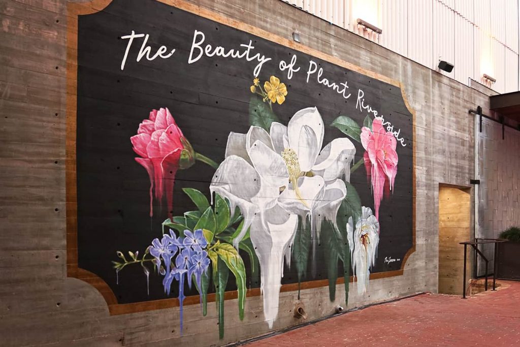 Mural with white, pink, and purple flowers painted on a black background with script that reads The Beauty of Plant Riverside