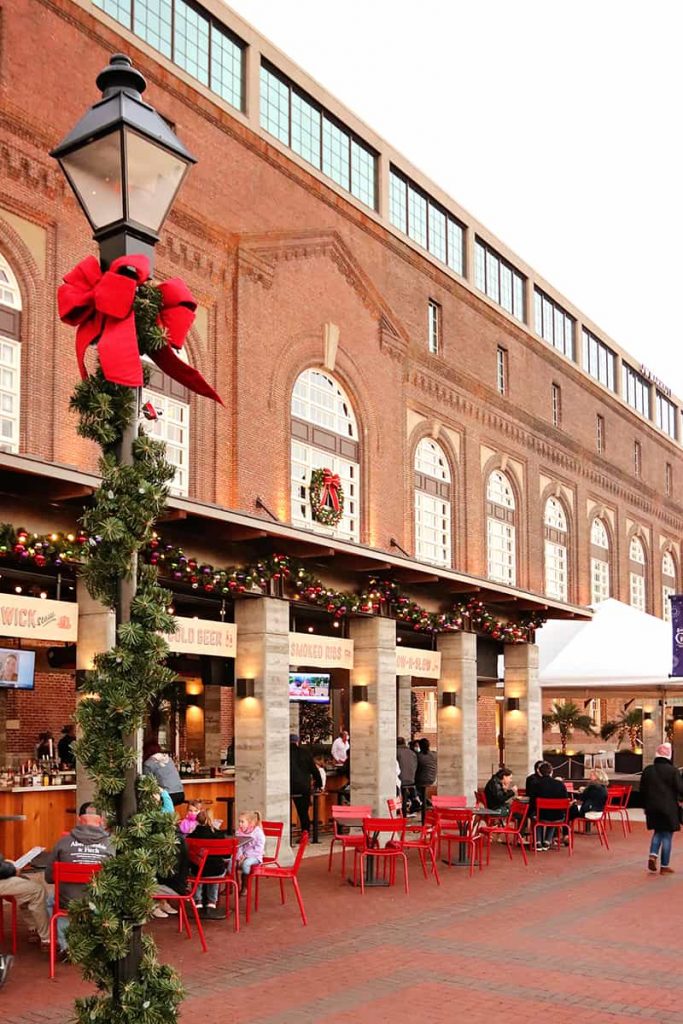 A lantern wrapped in holiday greenery and red ribbons in the foreground with a large historic brick building decorated with Christmas wreaths in the background