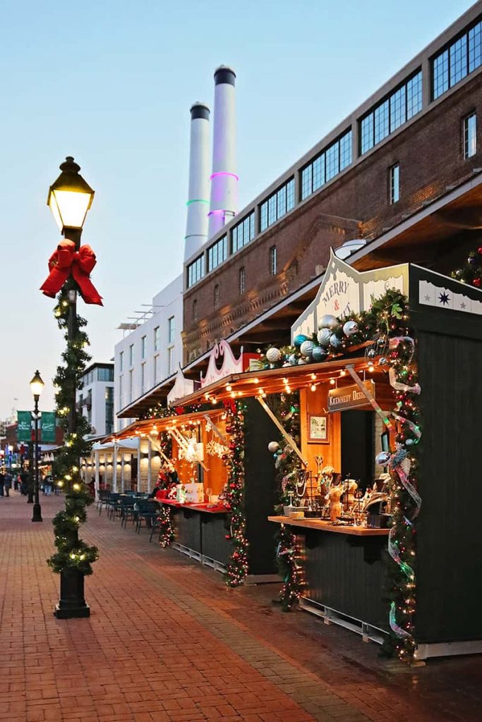 Christmas booths along the riverfront decorated with greenery and ornaments with the smokestacks of the JW Marriott Plant Riverside visible in the background