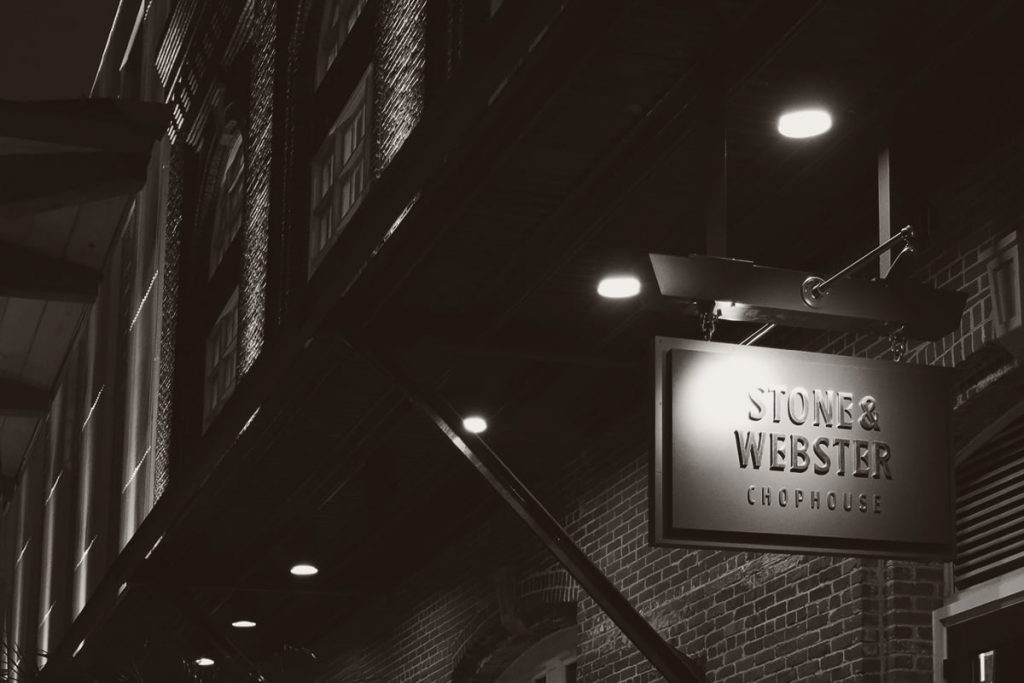 B&W image of a brick building at night with an illuminated sign for Stone & Webster Chophouse
