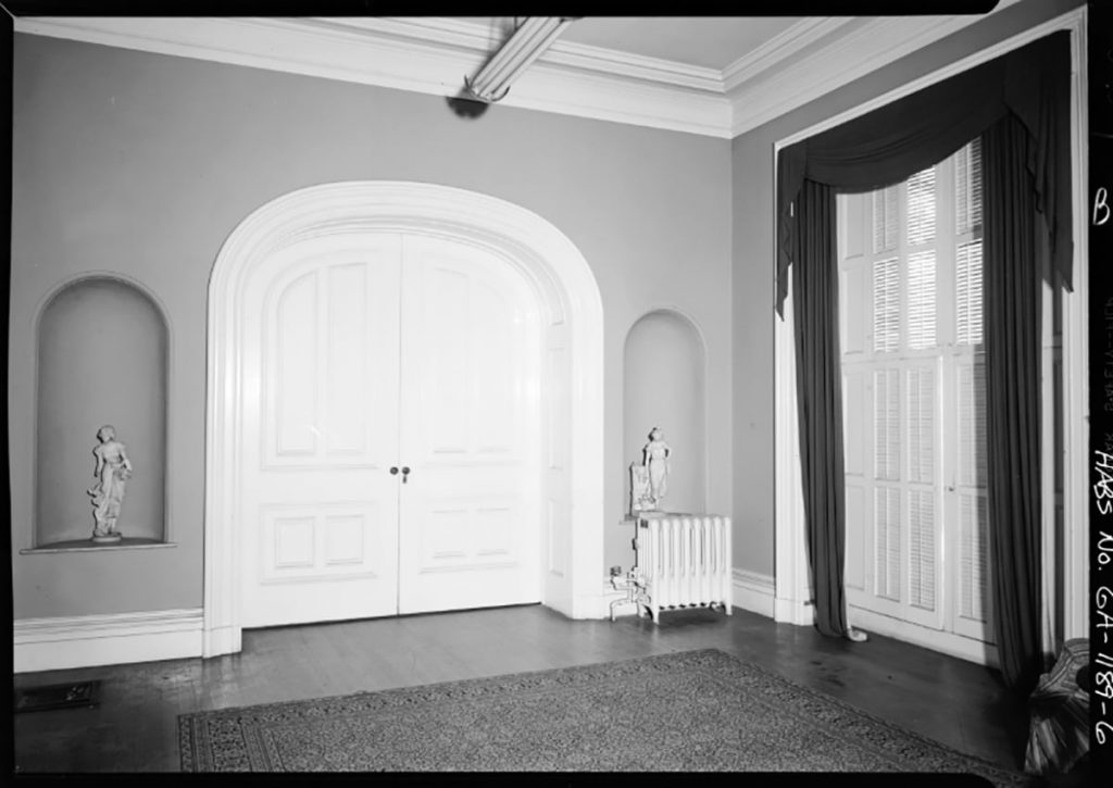Historic B&W image of the interior of Mercer House with curved niches and an arched double doorway, plus floor to ceiling windows, hardwood floors, and a white radiator