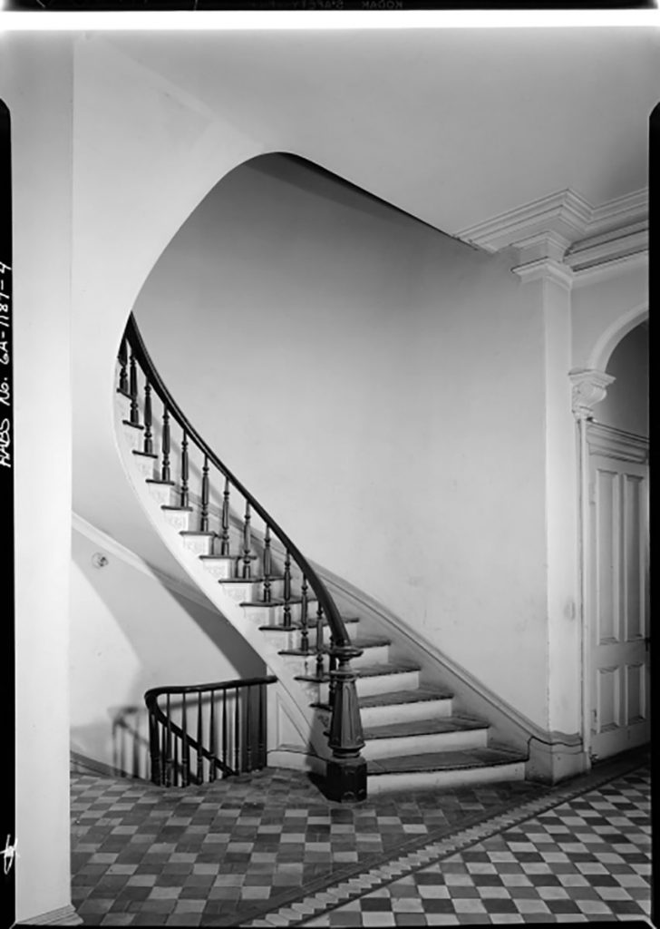Historic B&W image of the interior of the Mercer House showing an elegant curved staircase and parquet flooring in an intricate pattern