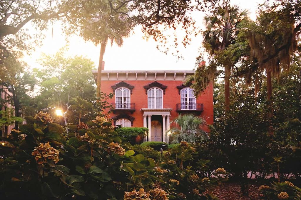 View from a distance of the brick front facade of the Mercer Williams House as seen through dried hydrangea bushes and large oak trees