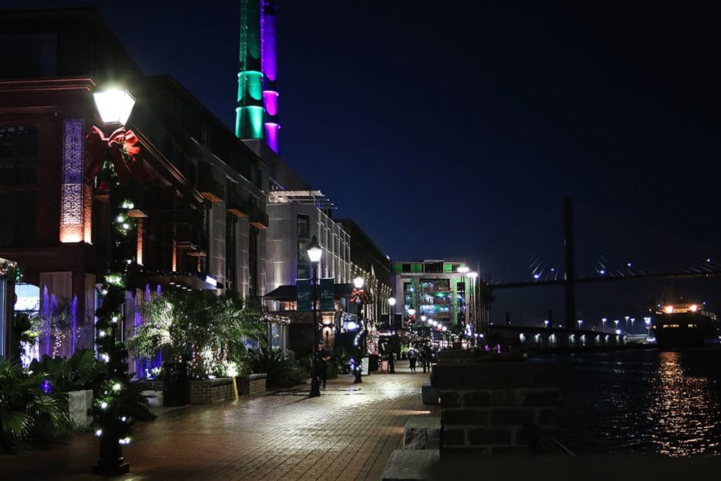 Nighttime shot of the Plant Riverside District showing the riverwalk, a large cargo ship in the Savannah River, and neon lights illuminating the shops and hotels along the riverfront