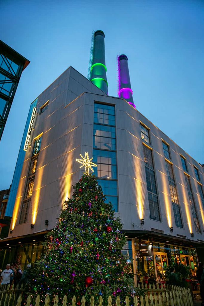 The two towers of the Plant Riverside District lit in purple and green for the holidays. A decorated Christmas tree stands below in the Savannah Christmas Market.