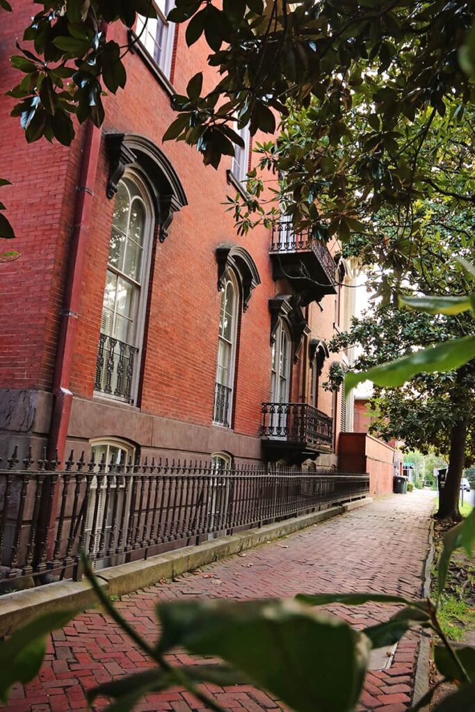 Side view of a 3-story brick home surrounded by a wrought-iron fence. The home has heavy iron details above the windows and features wrought-iron balconies