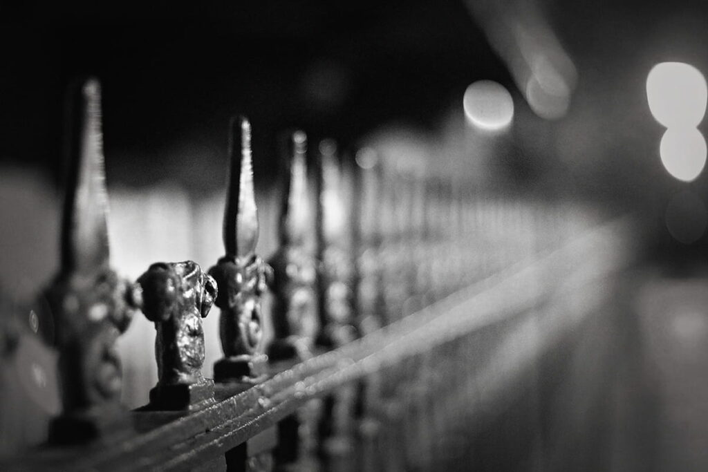 B&W image of the spikes on a wrought-iron fence with the tip of one spike broken and missing